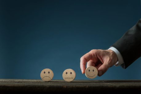 Hand of a businessman choosing a wooden cut circle with happy face on it in a conceptual image of satisfaction. Over blue background with copy space.